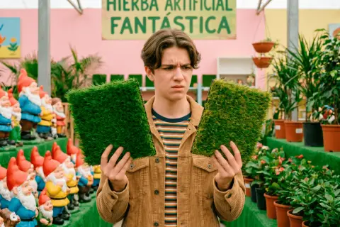 Joven indeciso compara dos muestras de césped artificial, buscando el mejor césped artificial del mercado. Detrás, gnomos de jardín y plantas.