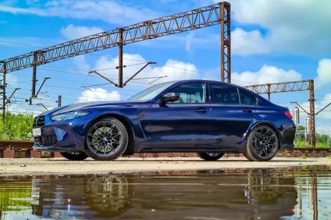 Granatowe BMW M3 G80 stoi obok torów kolejowych, odbijając się w kałuży.