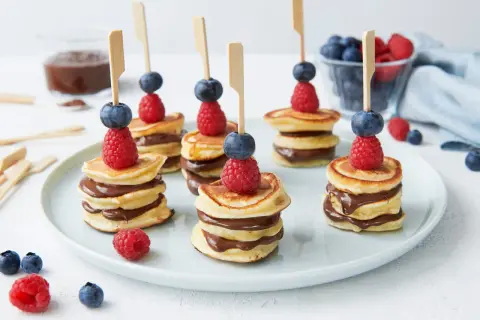 Mini pancakes rezept: gestapelte Mini-Pfannkuchen mit Schokocreme, garniert mit Himbeeren und Blaubeeren auf Spießen.