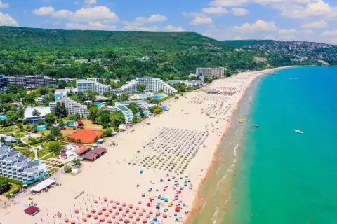 Słoneczne bułgarskie plaże tętnią życiem. Widać hotele, mnóstwo leżaków i parasoli, a także turystów kąpiących się w turkusowym morzu.