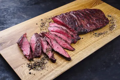 Perfekt zubereitetes Skirt Steak, in Scheiben geschnitten, liegt auf einem Holzbrett. Gewürze und Pfeffer sind verstreut.