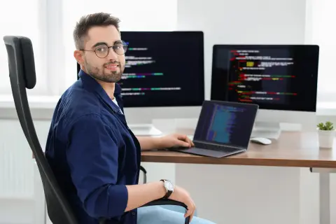 Typowy programista w okularach siedzi przy biurku z trzema monitorami wyświetlającymi kod.