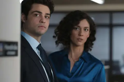 Besetzung von The Recruit: Ein Mann im Anzug und eine Frau in einem blauen Hemd blicken ernst in die Kamera.
