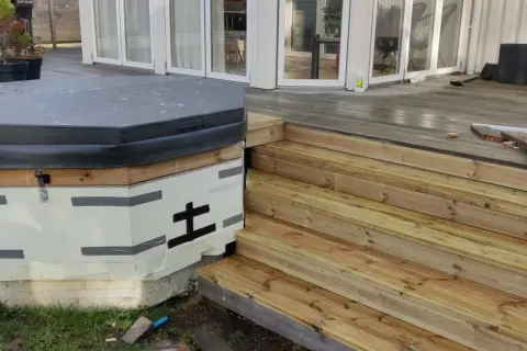 Pågår bygga badtunna. Isolerad stomme med trädetaljer och en grå presenning. Nya trätrappor leder upp till altanen.