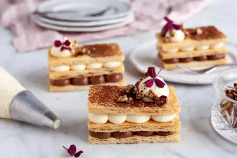 Tre eleganta lager av smördeg, chokladkräm och vaniljkräm toppas med nötter och en lila blomma. En lyxig dessert redo att avnjutas.