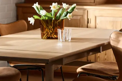 Ett ljust träbord med klassiska designstolar i läder. En vas med vita blommor och glas står på bordet.