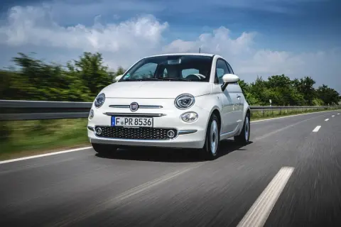 Biały fiat mini pędzi po asfaltowej drodze, otoczony zielenią i błękitnym niebem z chmurami.