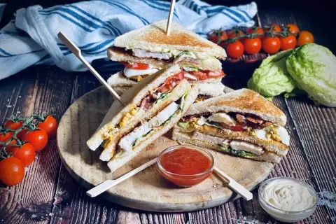 Leckere Sandwich-Ideen: Gestapelte Club-Sandwiches mit Hähnchen, Speck, Ei und Tomaten auf einem Holzbrett, daneben Tomaten und Salat.