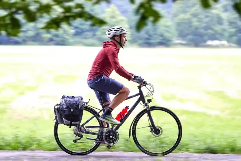Mężczyzna w kasku jedzie na rowerze trekkingowym z sakwami, ciesząc się przejażdżką pośród zieleni.