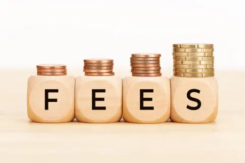 Cztery drewniane kostki z literami F, E, E, S tworzą napis FEES. Nad nimi piętrzą się monety, symbolizując rosnące opłaty.