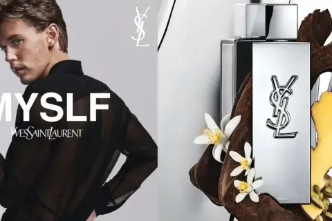 Młody mężczyzna w czarnej, prześwitującej koszuli, obok flakonu perfum Yves Saint Laurent MYSLF, otoczony drewnem i kwiatami.