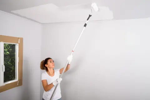Mujer pintando el techo con rodillo. La pregunta de si que se pinta primero el techo o las paredes es clave en esta tarea.