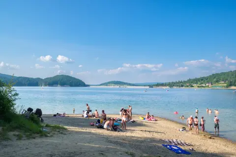 Słoneczny dzień na plaży w Polańczyku-zdroju. Ludzie odpoczywają nad wodą, niektórzy kąpią się, inni opalają.