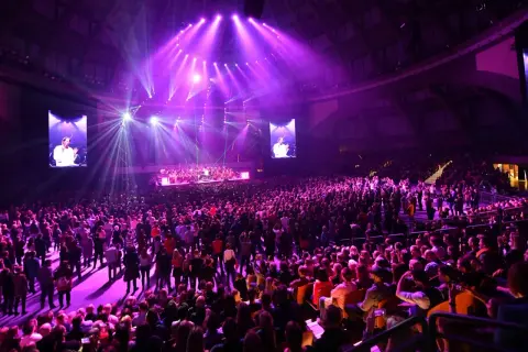 Wielki koncert w Stadionie Wrocław. Tłumy fanów, scena z orkiestrą i ekranami, na których widać dyrygenta.