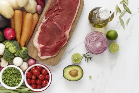 Un corte de carne magra, rodeado de verduras frescas como zanahorias, brócoli, tomates y aguacate, junto a aceite de oliva y limón.