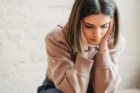 Een jonge vrouw met haar handen tegen haar slapen, diep in gedachten verzonken. Ze lijkt te worstelen met de gevolgen van gaslighting door vrouwen, haar blik neergeslagen.