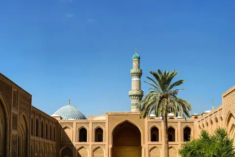 Ein Minarett und eine Kuppel einer Moschee ragen über traditionelle Gebäude mit Bögen und Palmen. Einblick in die reiche irakische Kultur.