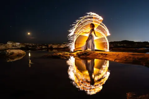 Une femme en robe blanche danse sous un ciel étoilé, entourée d'arcs de lumière incandescente. Le reflet de cette scène magique se mire dans l'eau calme, évoquant un coucher du soleil féerique.