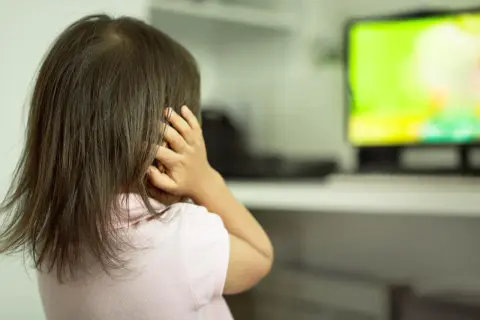 Dziewczynka z autyzmem zasłania uszy, reagując na bodźce. Ekran telewizora emituje jasne, intensywne kolory.