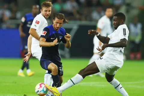 Piłkarze walczą o piłkę na boisku, jeden z nich w białej koszulce z logo Bundesligi, być może to przyszli bundesliga mistrzowie.