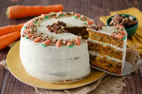 Torta di carote con glassa bianca, decorata con carote di zucchero e noci. Una fetta mostra gli strati soffici, perfetta per una carrot cake ricetta semplice.
