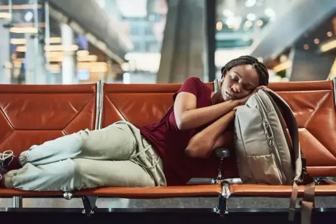 Frau schläft auf einer Bank im Flughafen bei Nacht, ihr Kopf ruht auf ihrem Rucksack.