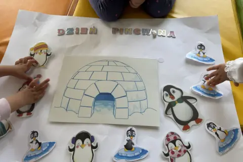 Dzieci tworzą plakat na dzień pingwina w przedszkolu, ozdabiając go naklejkami pingwinów i igloo.