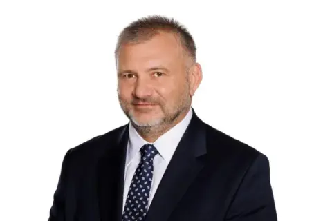 Mężczyzna w garniturze i krawacie, z krótkimi włosami i zarostem. Wygląda jak prokurator generalny.