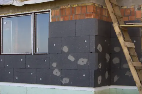 Budowa domu w toku, trwa ocieplanie ścian zewnętrznych styropianem. Widać okna i fragment rusztowania.