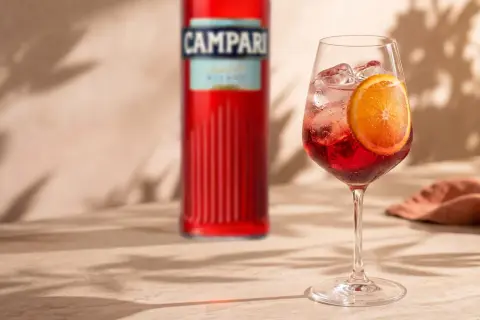 Orzeźwiający Campari Spritz z plasterkiem pomarańczy i kostkami lodu, obok butelki Campari. Idealny na letnie popołudnie.