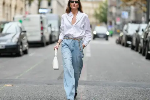Kobieta w luźnych, jasnych jeansach typu mom fit i białej koszuli, z białą torebką. Idealny look dla każdej kobiety ceniącej wygodę i styl.