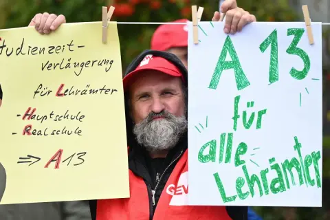 Mann mit Bart und roter Weste hält Schilder hoch. Ein Schild fordert "A13 für alle Lehrer" und thematisiert das Lehrer Gehalt Berlin.
