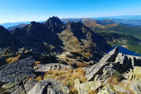Widok z kamienistego szczytu w Tatrach Wysokich na skaliste granie, trawiaste zbocza i turkusowe jezioro.