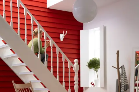 Ein rotes, holzvertäfeltes Treppenhaus mit weißer Balustrade. Eine Frau sitzt auf der Treppe. Ein Hirschkopf und ein Baum schmücken die Wand. So lässt sich ein Treppenhaus gestalten.