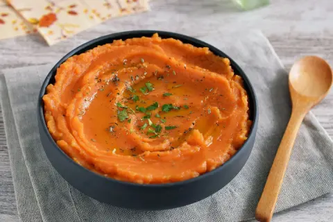 Un tazón de puré de boniato gourmet, cremoso y adornado con aceite y hierbas. Una cuchara de madera descansa a su lado.