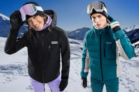 Pareja sonriente en la nieve, ella con chaqueta hardshell negra, él con chaqueta acolchada verde.