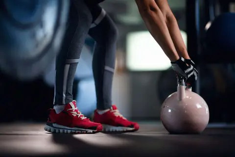 Person mit roten Schuhen und Trainingshandschuhen hebt abends eine Kettlebell für das Krafttraining.