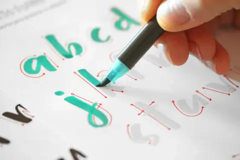 Mano practicando lettering con plantillas imprimibles gratis. Letras "a, b, c, j, i, t, u" en verde y rojo.