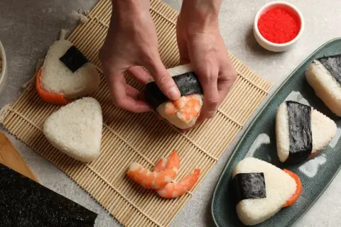 Ręce formują onigiri z krewetkami, tworząc pyszne japońskie jedzenie. Obok miska ryżu i talerz z gotowymi przysmakami.