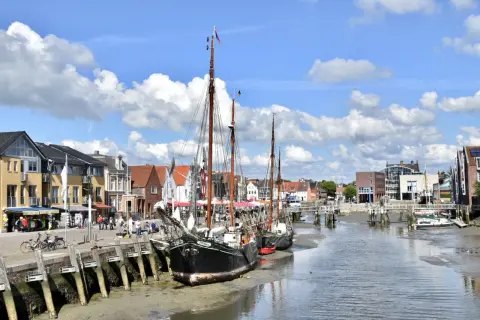 Tre segelfartyg ligger vid kaj i Husum, Tyskland. Bakom dem syns en rad hus och en bro över kanalen.