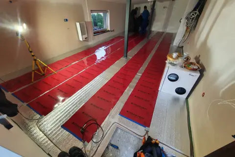 Instalacja folii grzewczej. Widać długie pasy czerwonej folii na podłodze, przygotowane do montażu. Wady tej metody mogą obejmować nierównomierne rozprowadzanie ciepła.