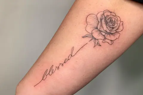 Ein zarter Fineline-Schriftzug "blessed" mit einer kleinen Rose als Tattoo.