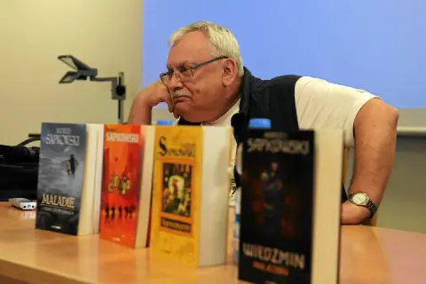 Mężczyzna zamyślony nad stosem książek, wśród nich "Wiedźmin" i inne tytuły Sapkowskiego, idealne dla fanów szukających książek podobnych do Wiedźmina.