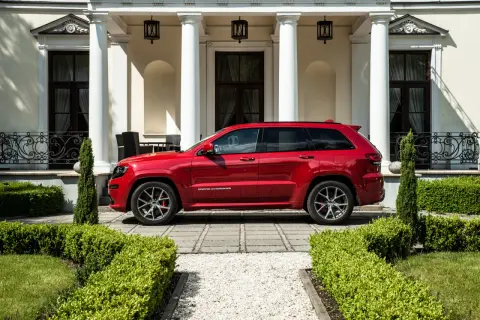 Czerwony Jeep Grand Cherokee SRT zaparkowany przed elegancką posiadłością z białymi kolumnami i zadbanym ogrodem.