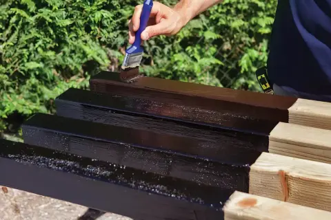 Mano che applica vernice scura su listelli di legno grezzo, un passo essenziale per imparare come impermeabilizzare il legno e proteggerlo.
