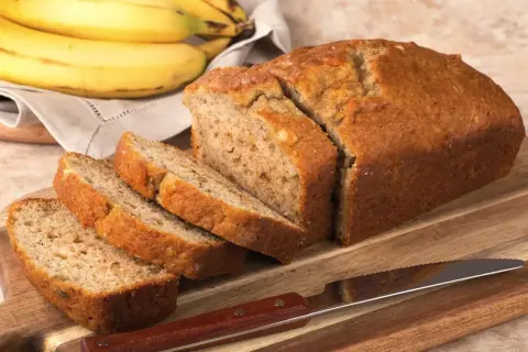 Chlebek bananowy, pokrojony na kromki, leży na desce obok noża. Obok pęk dojrzałych bananów. Kwestia smaku, czy wolisz go na ciepło czy na zimno.