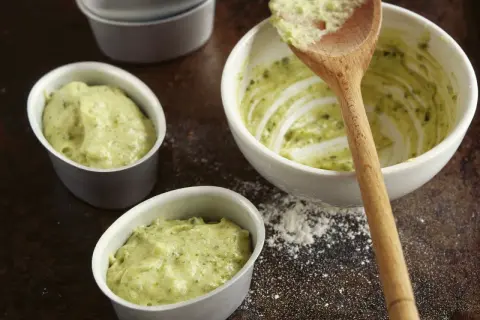 Kleine Förmchen werden mit einer grünen Masse gefüllt, die nach Pistaziencreme selber machen aussieht. Ein Holzlöffel liegt in einer Schüssel mit der Masse.