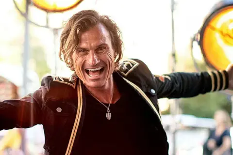 Petter Stordalen, trots sin ålder, strålar av energi i en mörk jacka med gulddetaljer. Han ler brett och gestikulerar.