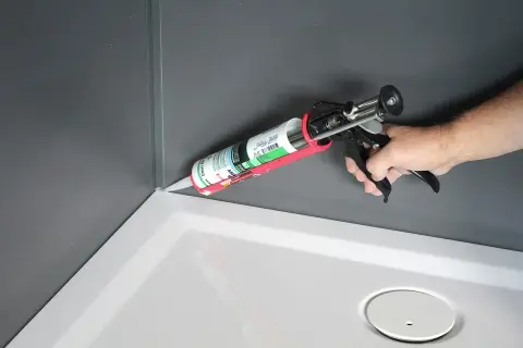 Aplikacja silikonu do kabiny prysznicowej. Dłoń trzyma pistolet z tubą silikonu, uszczelniając narożnik brodzika.