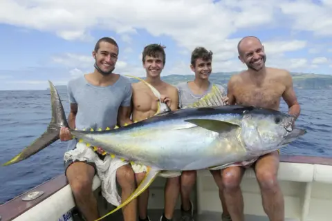 Vier Männer präsentieren stolz einen riesigen Thunfisch auf einem Boot vor den Azoren. Ein erfolgreicher Tag beim **Azoren Angeln**!